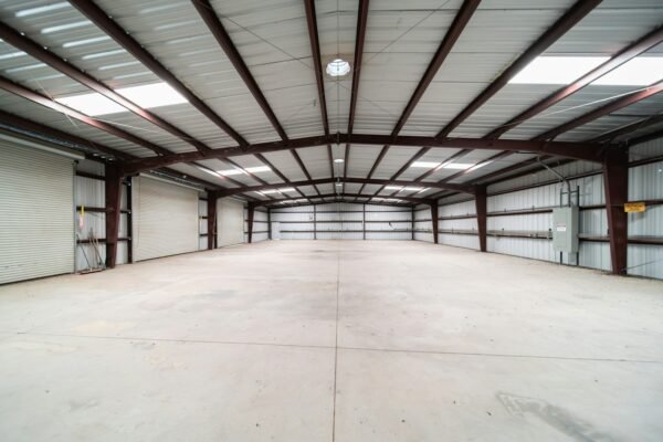 Empty steel-framed warehouse interior with roller doors and a bare concrete floor, lit by skylights, no people present