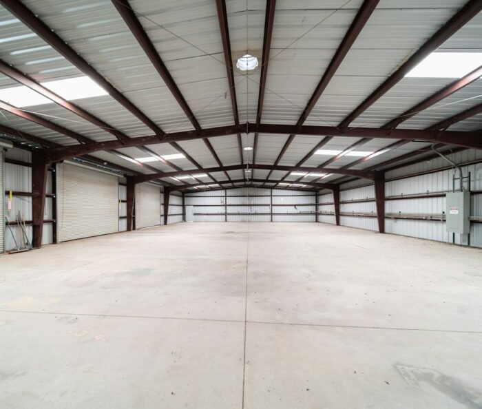 Empty steel-framed warehouse interior with roller doors and a bare concrete floor, lit by skylights, no people present
