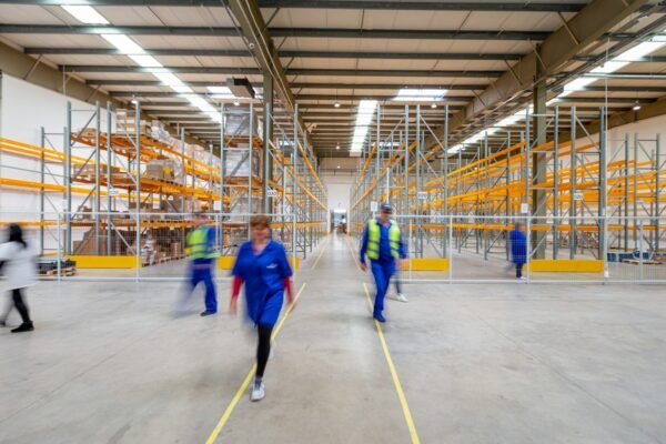 Workers in hi-vis vests walking through a wide concrete aisle between tall steel warehouse storage racking
