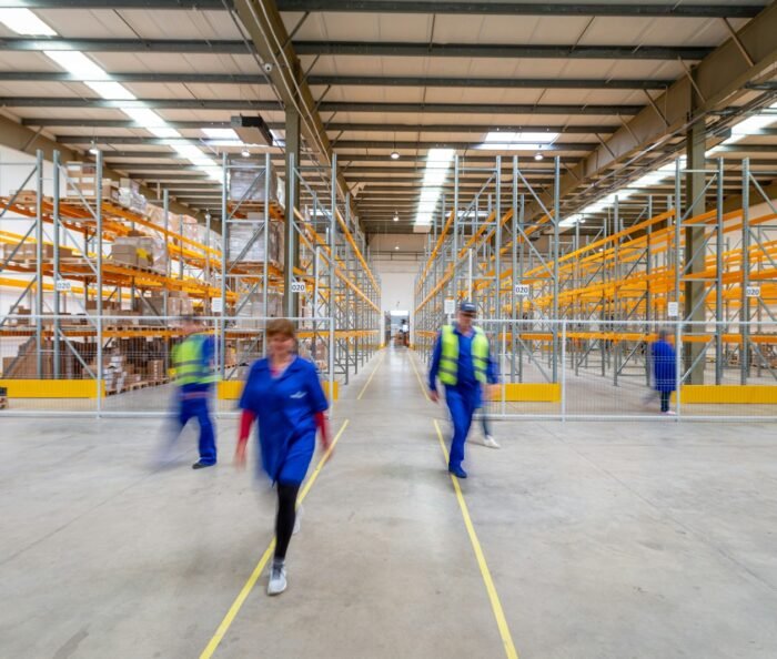 Workers in hi-vis vests walking through a wide concrete aisle between tall steel warehouse storage racking