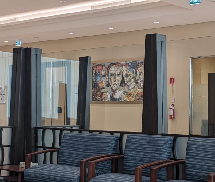 Empty modern clinic waiting area with rows of blue upholstered chairs, dark glass partitions and a framed abstract mural.