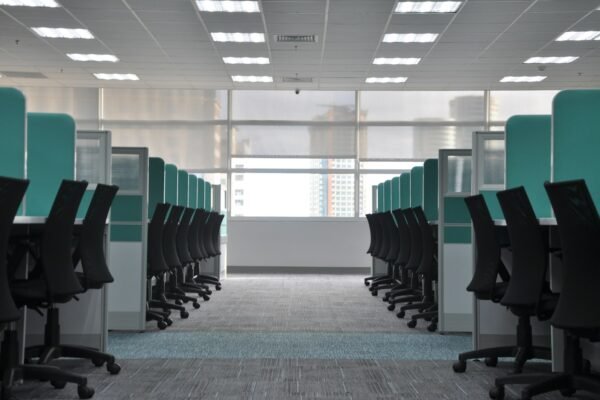 Rows of empty black office chairs and teal cubicle dividers in a vacant open-plan office with a soft city view