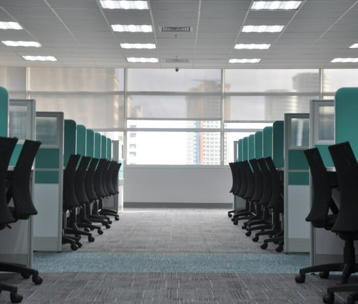 Rows of empty black office chairs and teal cubicle dividers in a vacant open-plan office with a soft city view