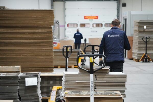 Warehouse worker seen from behind pushing a loaded hand pallet jack past stacked materials in an industrial storage facility