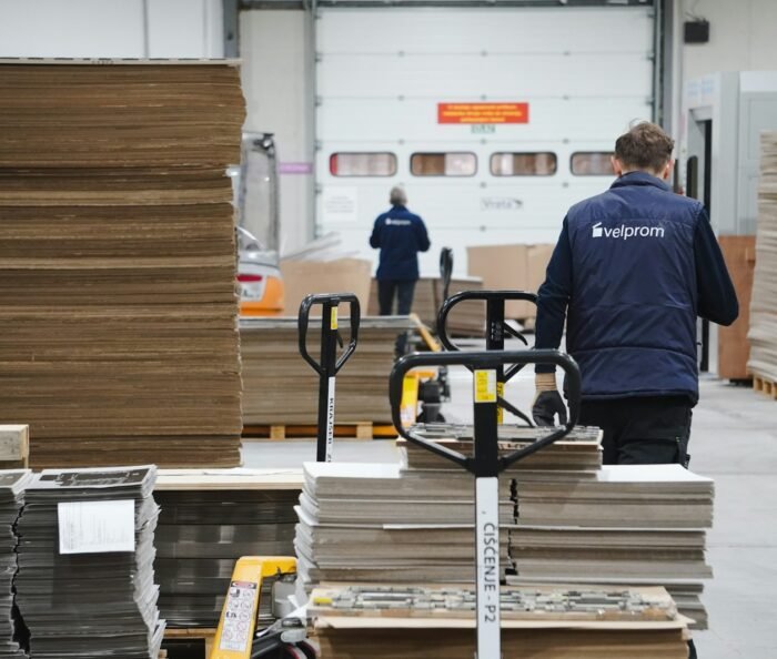 Warehouse worker seen from behind pushing a loaded hand pallet jack past stacked materials in an industrial storage facility