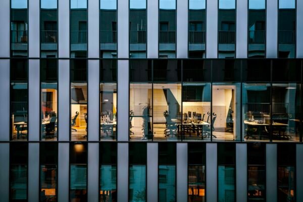 Empty desks and vacant office chairs in a lit workspace seen through a cool blue glass facade at dusk