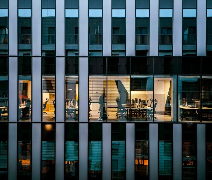 Empty desks and vacant office chairs in a lit workspace seen through a cool blue glass facade at dusk