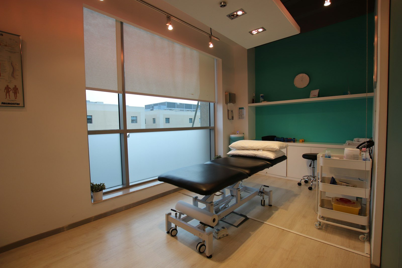 Physiotherapy treatment table in a clinic room with a teal feature wall and small hand weights on a shelf